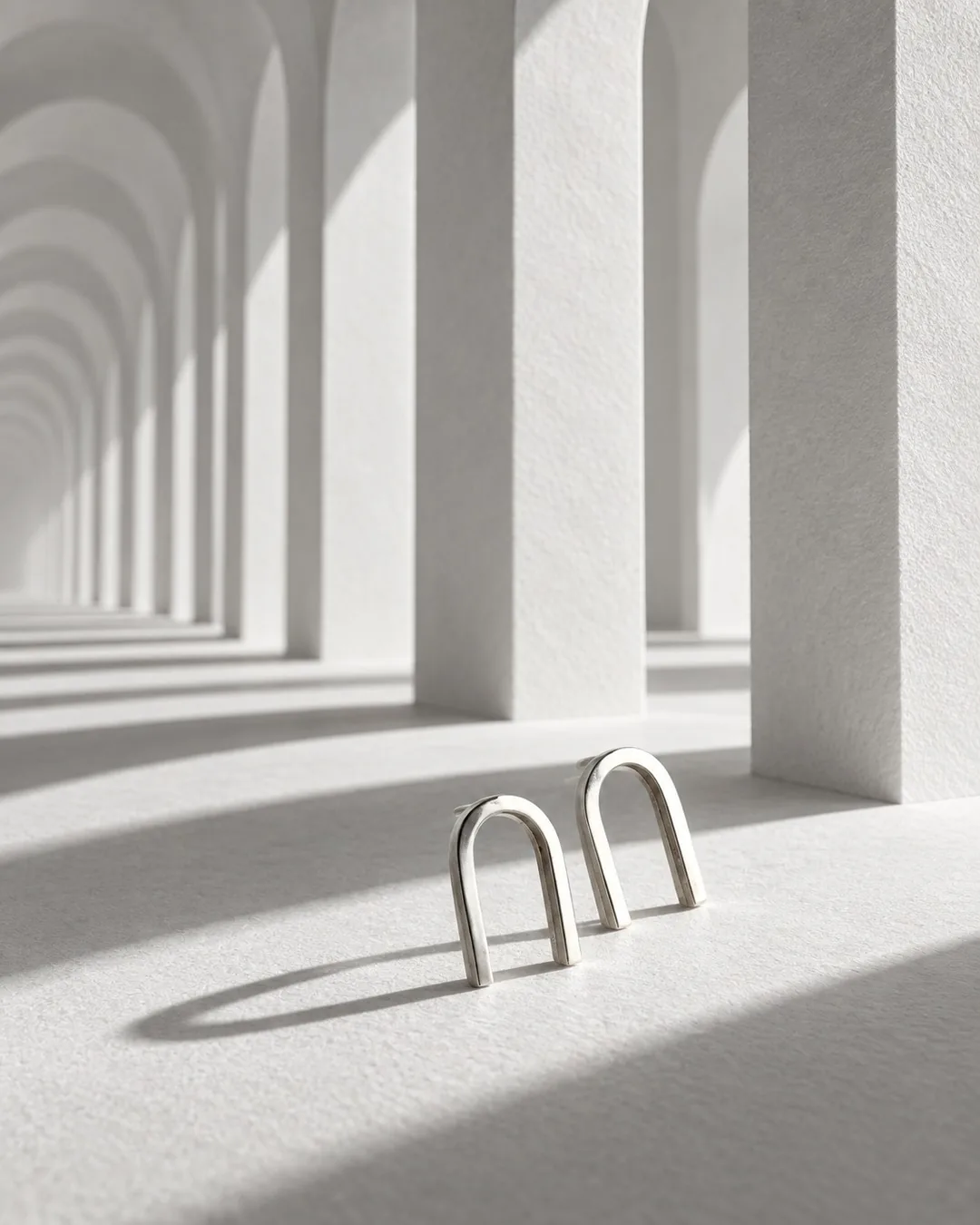 Brinco arco duplo de prata diante de uma colunata de arcos brancos, com sombras longas